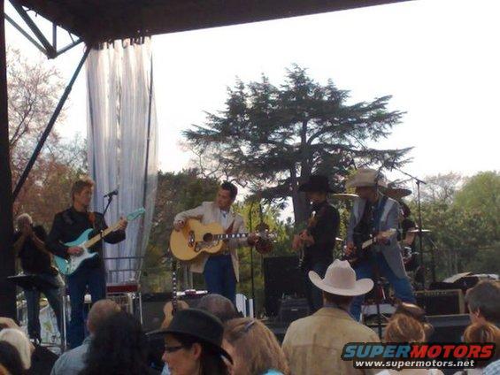 Band playing guitars and singing on a stage outdoors with trees in the background