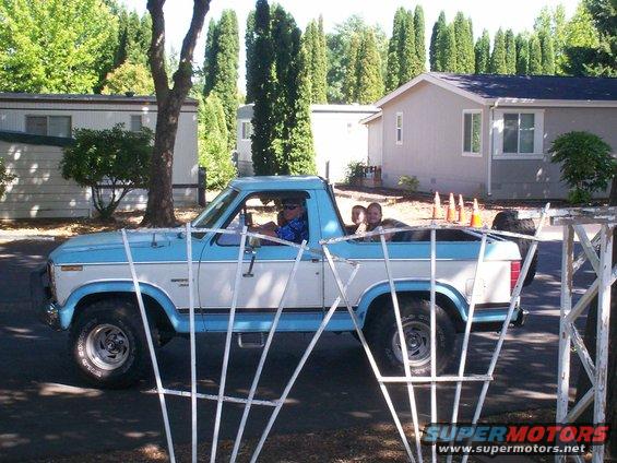 Light blue and white Ford pickup truck driving past mobile homes, with two children in the bed