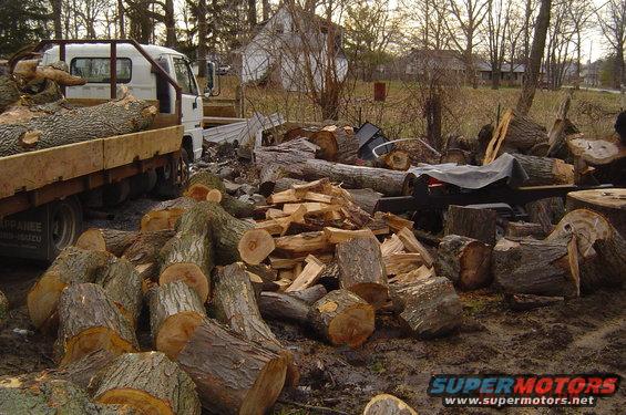 Flatbed truck loaded with logs, parked next to a pile of cut firewood