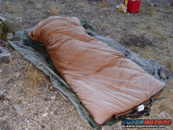 Orange sleeping bag on a camouflage tarp on dry ground. A red cooler is in the background
