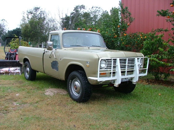 1975 International Scout II pickup truck with a white brush guard parked on grass, with a red barn and orange trees in the background