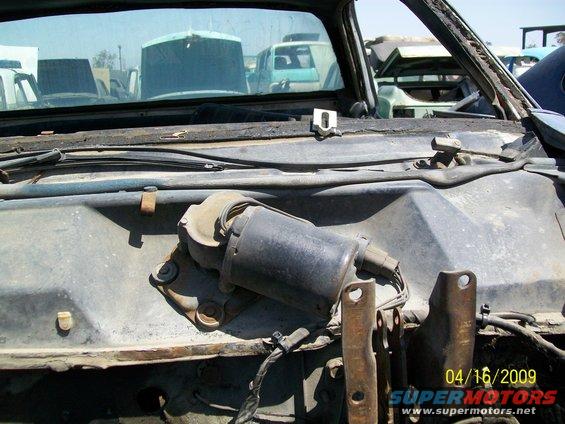 Worn windshield wiper motor assembly on a dusty dashboard with other vehicles in the background