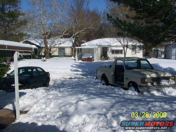Ford pickup truck stuck in snow, door open. Another car is partially visible on the left