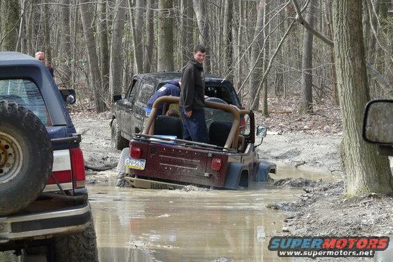 Several off-road vehicles are stuck in deep mud in a wooded area. One Jeep is partially submerged