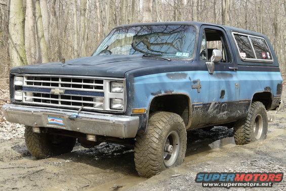 Mud-covered blue and black Chevrolet K5 Blazer with aftermarket hood, stuck in mud