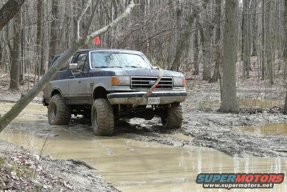 Ford Bronco with lifted suspension driving through a muddy forest trail. License plate reads 292W24