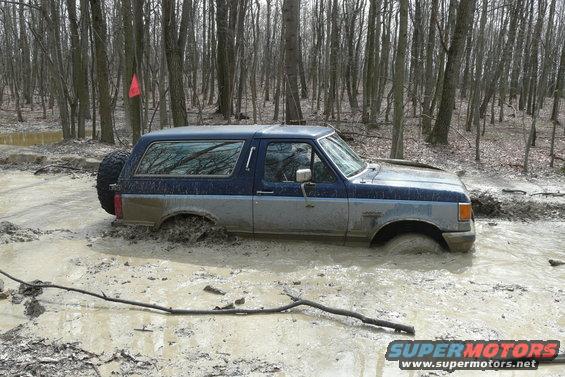 Two-tone blue and gray Ford Bronco stuck in mud, spare tire visible on rear
