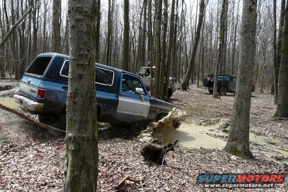 Blue and silver SUV stuck in mud, trees and other vehicles in background
