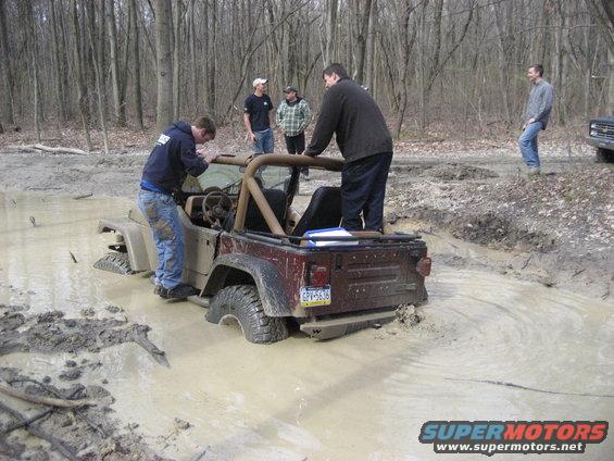Jeep Wrangler stuck in mud pit with several people attempting to help; another vehicle visible in background