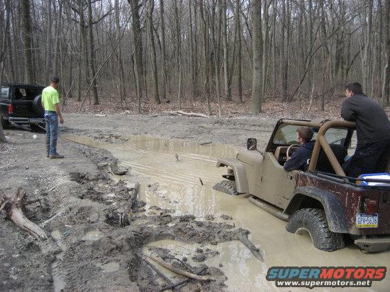 Tan Jeep Wrangler stuck in mud, with a black Ford Bronco in the background