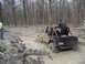 1991 Ford Bronco - A tan Jeep Wrangler is stuck in the mud while being pulled by a chain in a wooded area.