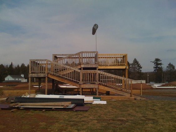 Wooden observation deck with ramps and railings, under a cloudy sky. Construction materials are piled in the foreground
