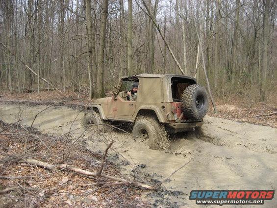 Tan Jeep Wrangler driving through a muddy trail in a forest, with a spare tire mounted on the back