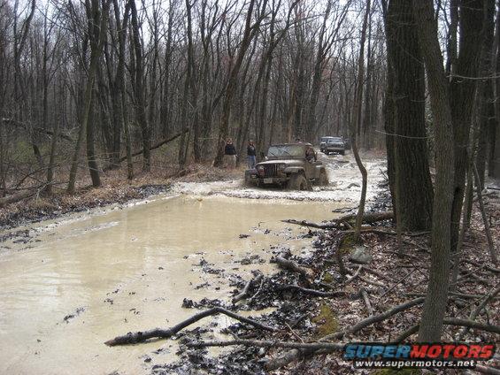Brown Jeep Wrangler driving through a muddy forest trail with another vehicle behind it