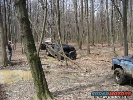 1991 Ford Bronco stuck in mud, spare tire visible on rear, in a wooded area