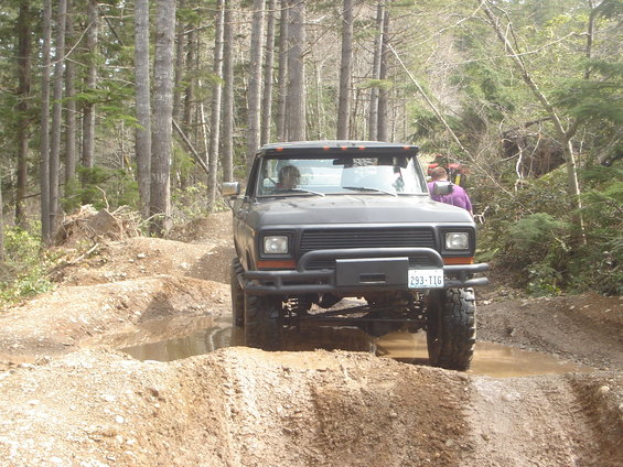 Dark gray Ford Bronco driving through muddy terrain with trees in the background