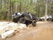 1989 Ford Bronco - A modified black Ford Bronco navigates a rocky obstacle course in a wooded area, with other vehicles nearby.