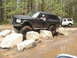 1989 Ford Bronco - A black Ford Bronco is shown off-roading over large rocks, with another white Bronco visible in the background.