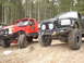1989 Ford Bronco - Two off-road trucks, a red one and a black one, are parked side-by-side on a gravelly path surrounded by trees.