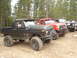1989 Ford Bronco - A line of off-road vehicles, including a black SUV with large tires, parked on a dirt road with trees in the background.