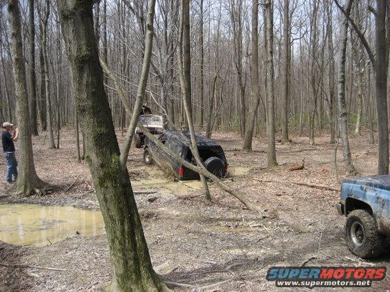 Two off-road vehicles stuck in mud in a wooded area. One is tilted and partially submerged