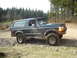 1989 Ford Bronco - This dark green Ford Bronco is shown off-road on a rocky path, with trees in the background.