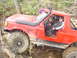 1989 Ford Bronco - Red 1989 Ford Bronco off-roading through mud and rocks with occupants inside.