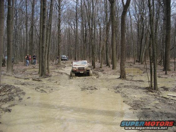 Mud-splattered Jeep Wrangler driving through a muddy forest path, front view