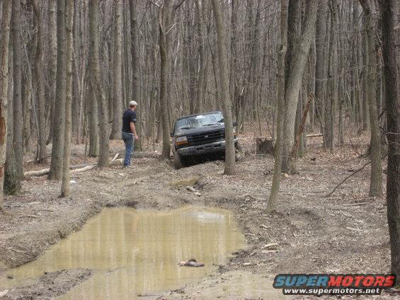 Man standing by a black Ford Bronco stuck in a muddy trail in the woods
