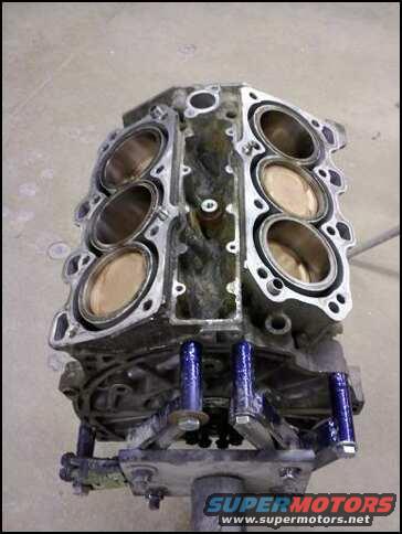 Engine block with cylinders visible, mounted on a stand. Supermotors.net logo at bottom
