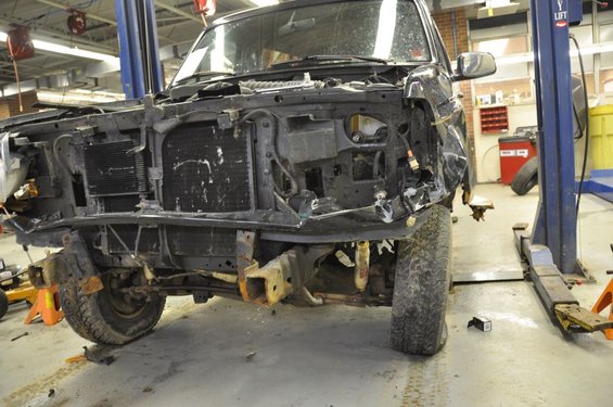 SUV front end is torn apart, missing grille and bumper, on a lift in a garage