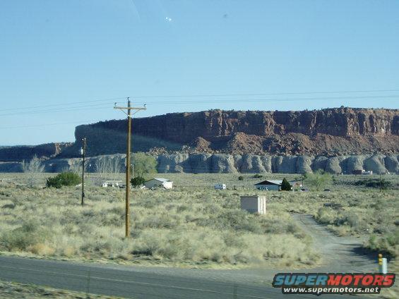 Desert landscape with mesas, houses, and telephone poles under a clear blue sky. A dirt road leads to a house