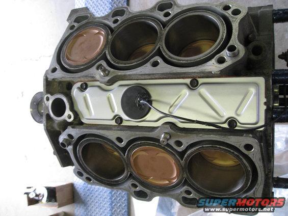 Engine block with six cylinders, copper-topped pistons visible in four cylinders. A wire extends from a black circular fitting