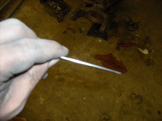 Hand holding a thin metal rod over a concrete floor with disassembled car parts and a red rag