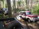 1979 Ford Bronco - Two off-road vehicles navigate a wet, wooded path, kicking up mud as they go.
