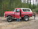 1979 Ford Bronco - Red Ford Bronco with a white top and lifted suspension parked on a dirt road in a wooded area.