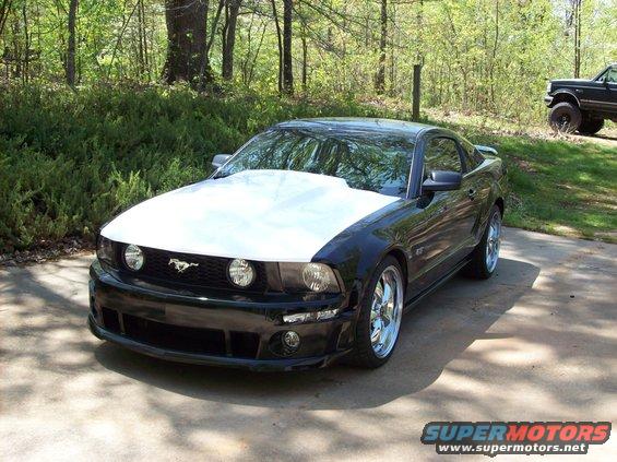 Black 2007 Ford Mustang coupe with a white hood, parked on a driveway. Another vehicle is visible in the background