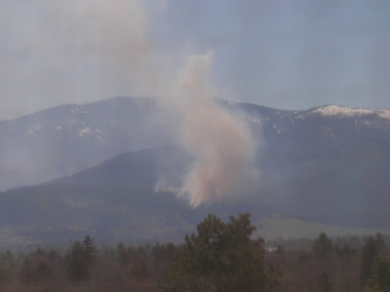 Smoke plume rising from a mountainside with snow-capped peaks in the distance