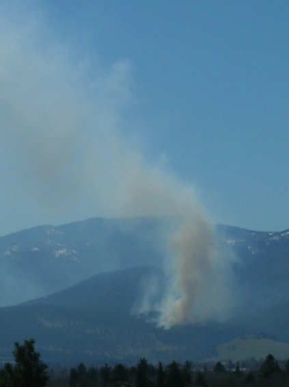 Smoke rising from a wildfire on a mountainside under a clear blue sky