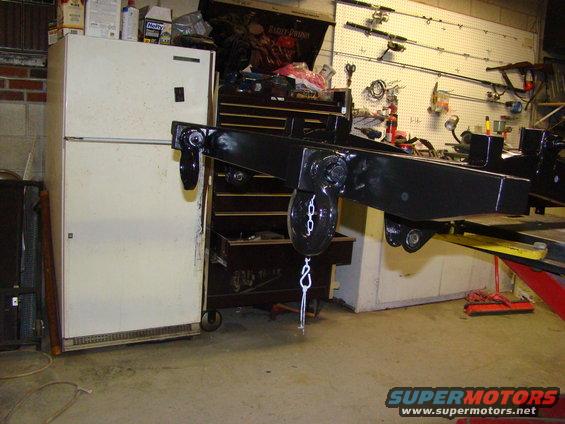 Black metal frame with pulley and chain hanging, next to a white refrigerator and tool chest