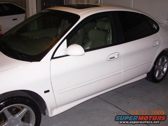 White sedan parked indoors, side view. Another white vehicle partially visible in background