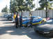 1995 Ford Probe - A group of people gather around a blue and yellow race car with its hood open, parked in a line with other vehicles.