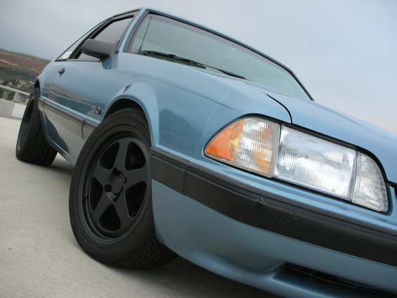 Light blue 1990 Ford Mustang with black five-spoke wheels, seen from a low front angle