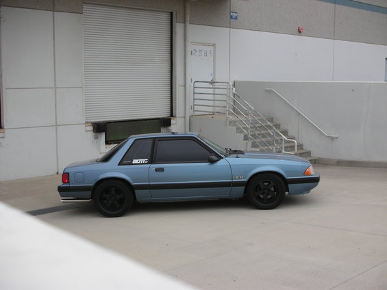 Blue 1990 Ford Mustang coupe with black wheels and a "BOTT" sticker on the rear quarter window, parked outside a warehouse