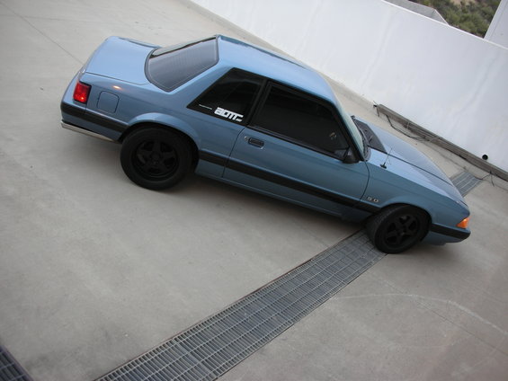 Blue 1990 Ford Mustang coupe with black wheels and a "BOTT" decal on the door, viewed from a high angle