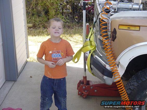 Brown and white truck lifted on a jack, front quarter view. Text on child's shirt reads 'HOW COOL AM I?'