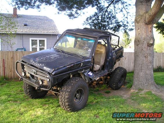 Black modified SUV with off-road tires, custom roll cage, and front bumper, parked on grass next to a tree