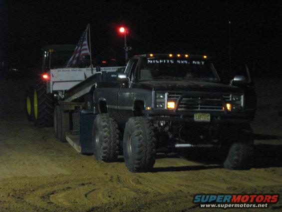 Modified pickup truck with large tires towing a tractor at night. Text on windshield reads 'MISFITS 4X4. NET'