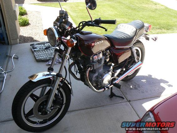 Maroon Honda motorcycle parked on concrete with a toolbox nearby. Part of a red car is visible on the right