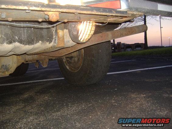 Underside view of a vehicle showing the rear tire, fuel tank, and exhaust system. The Supermotors logo is visible at the bottom right